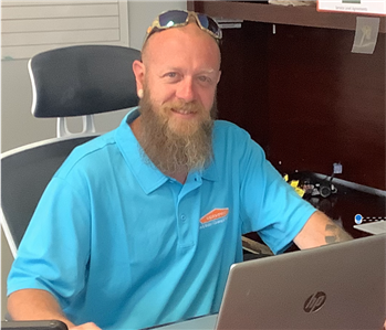 Bald middle-aged man, blue eyes, long beard, wearing a blue golf shirt with SERVPRO logo; seated at desk with laptop computer
